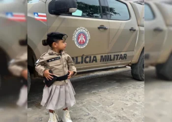 Liz Dorea, de 2 anos, sorridente vestindo uma farda improvisada da PMBA, em pé diante de uma viatura oficial em Castro Alves