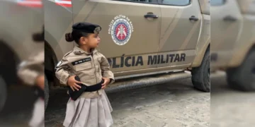Liz Dorea, de 2 anos, sorridente vestindo uma farda improvisada da PMBA, em pé diante de uma viatura oficial em Castro Alves